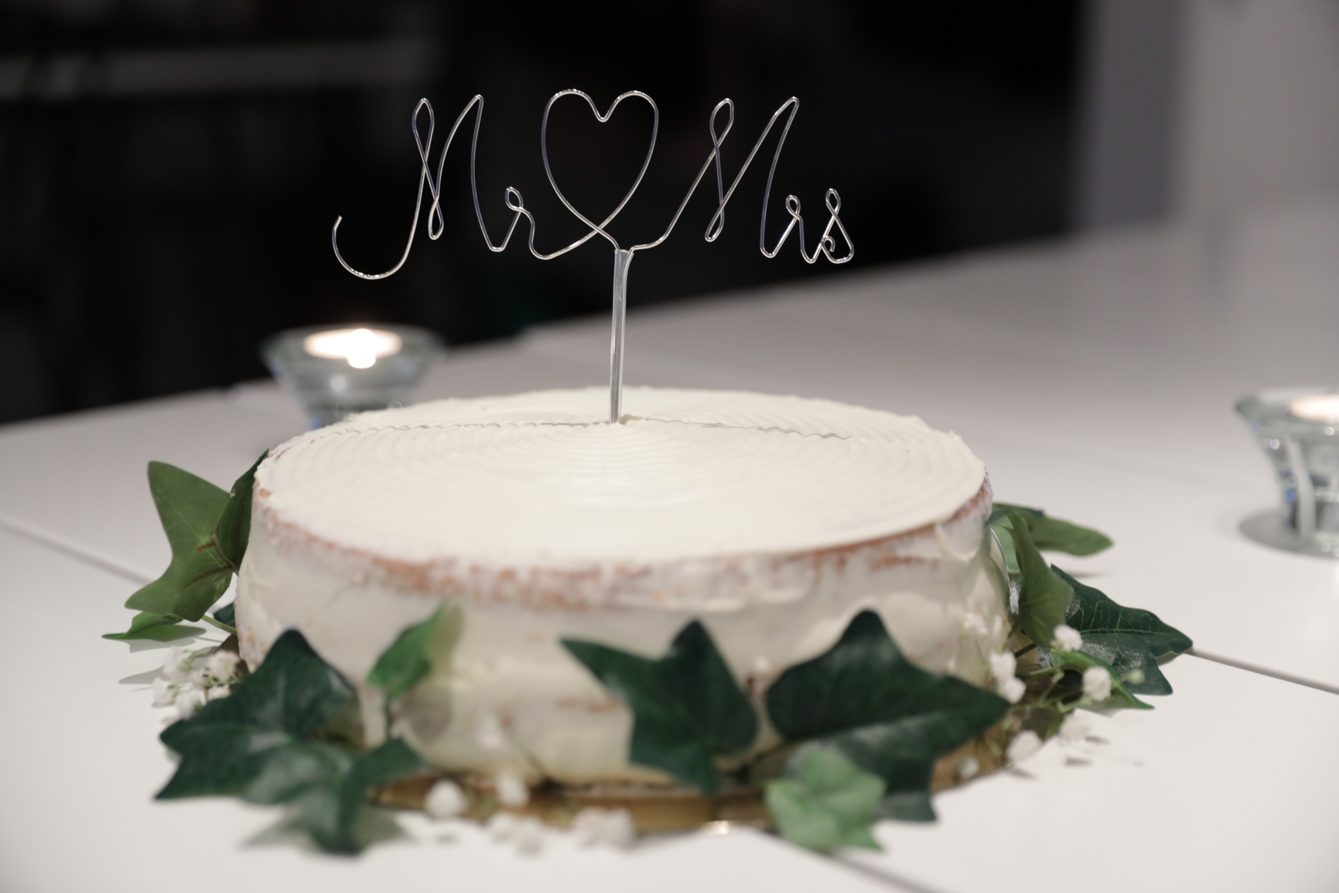 En vit bröllopstårta dekorerad med grönska och en toppdekoration i form av texten "Mr ♥ Mrs" i tråd. Tårtan står på ett vitt bord med tända ljus i bakgrunden.